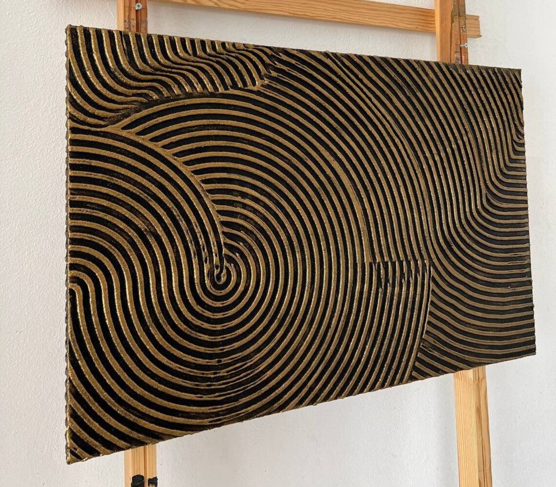 Metallic abstract canvas painting with gold and black concentric circular patterns.